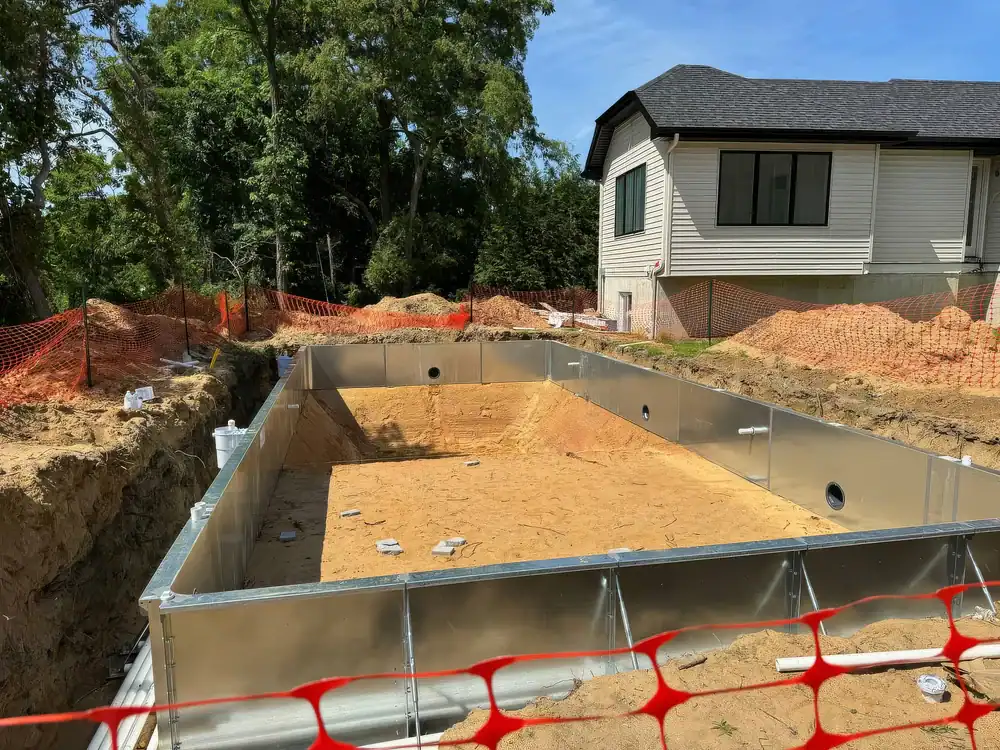 A rectangular in-ground pool under construction in a Douglas County, GA backyard, surrounded by sand, dirt mounds, and orange safety fencing, with a house and trees in the background.