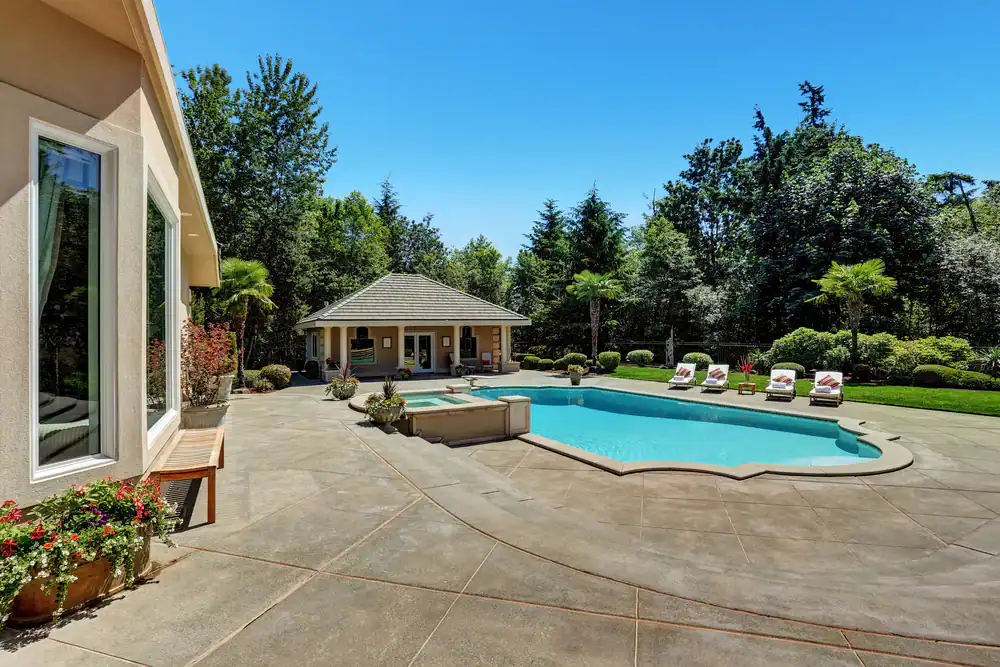 Spacious backyard in GA with an in-ground swimming pool crafted by expert pool construction Douglas County, several lounge chairs, a hot tub, a pool house, and lush trees and greenery under a clear blue sky.