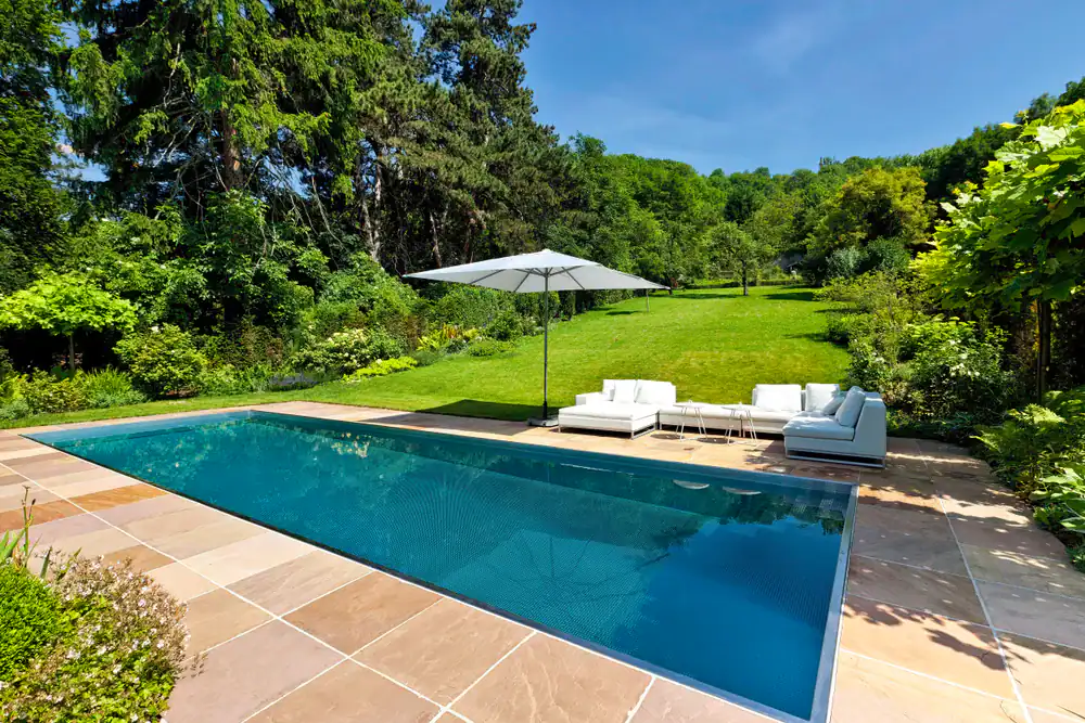A rectangular swimming pool with clear blue water, crafted by expert pool construction in Douglas County, GA, is surrounded by a stone patio. White outdoor sofas and a large umbrella sit beside the pool, framed by lush grass and trees under a clear sky.