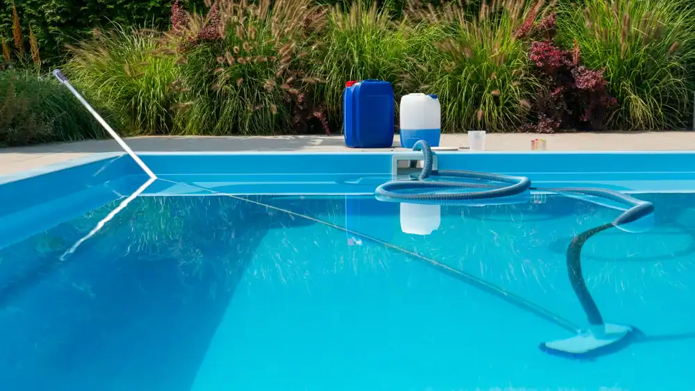 A swimming pool in GA is being cleaned with a pool vacuum and net. In the background, large containers of pool chemicals and supplies from a recent pool construction Douglas County project sit near tall grass and bushes by the poolside.