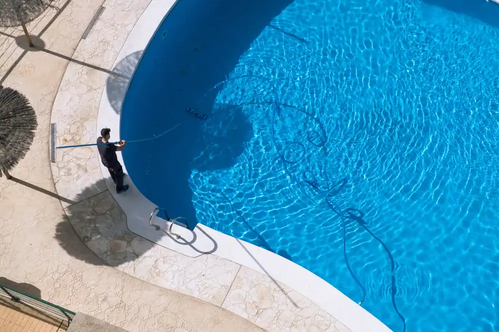 A person stands at the edge of an outdoor swimming pool, cleaning it with a long pole. The pool, built by pool construction experts in Douglas County, GA, is mostly empty, with clear blue water and cleaning equipment inside. Shadows fall across the deck.