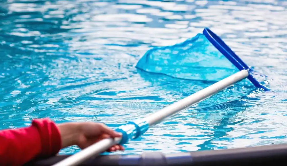 A person in GA uses a blue pool skimmer net to clean debris from the surface of clear blue pool water, showcasing the results of quality pool construction Douglas County residents trust.