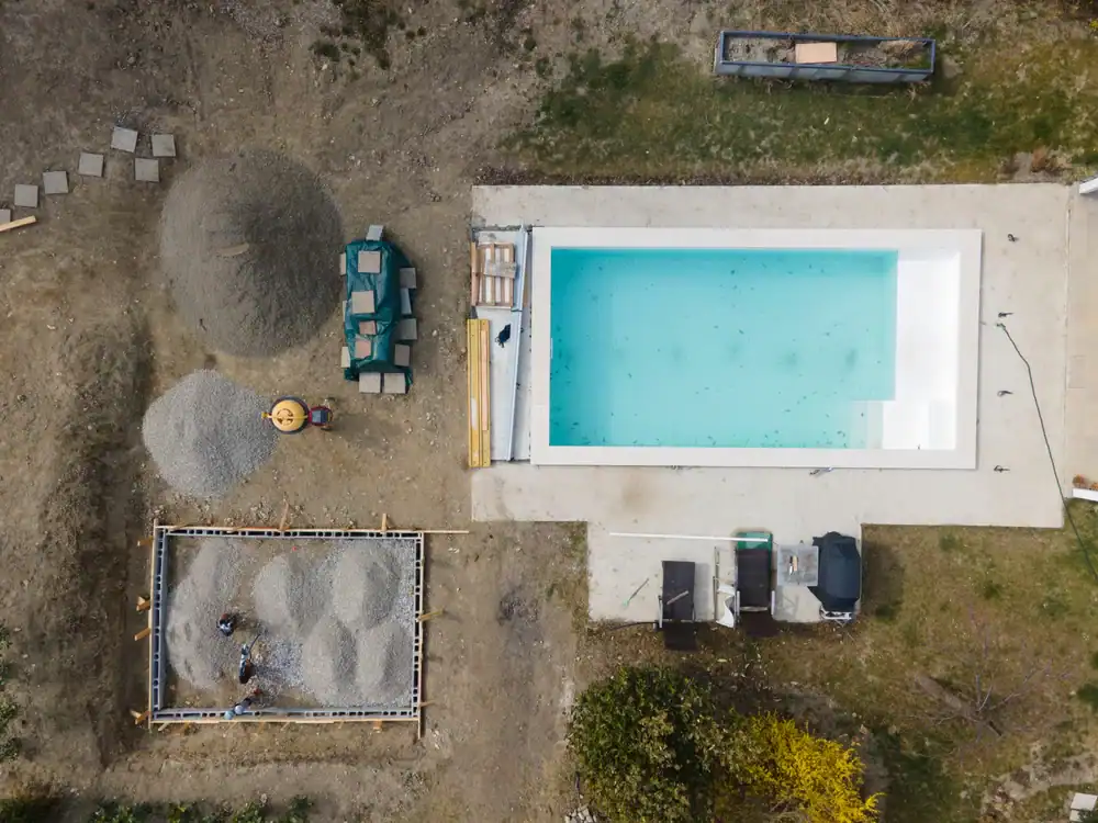 Aerial view of pool construction in Douglas County, GA, showing a rectangular swimming pool, construction materials, gravel piles, a wheelbarrow, and people working on a square frame in a backyard area.