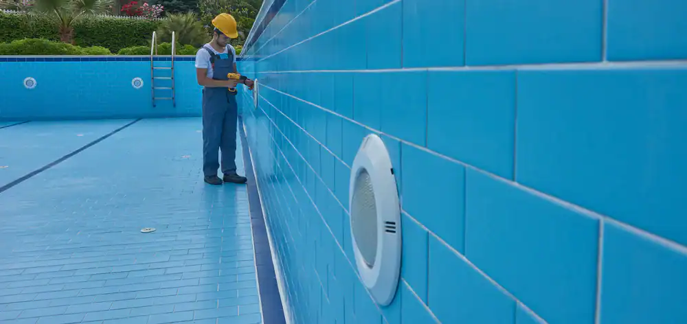 A worker in a yellow hard hat and blue overalls uses a power tool inside an empty, blue-tiled swimming pool during pool construction Douglas County, GA. A pool ladder and greenery are visible in the background.
