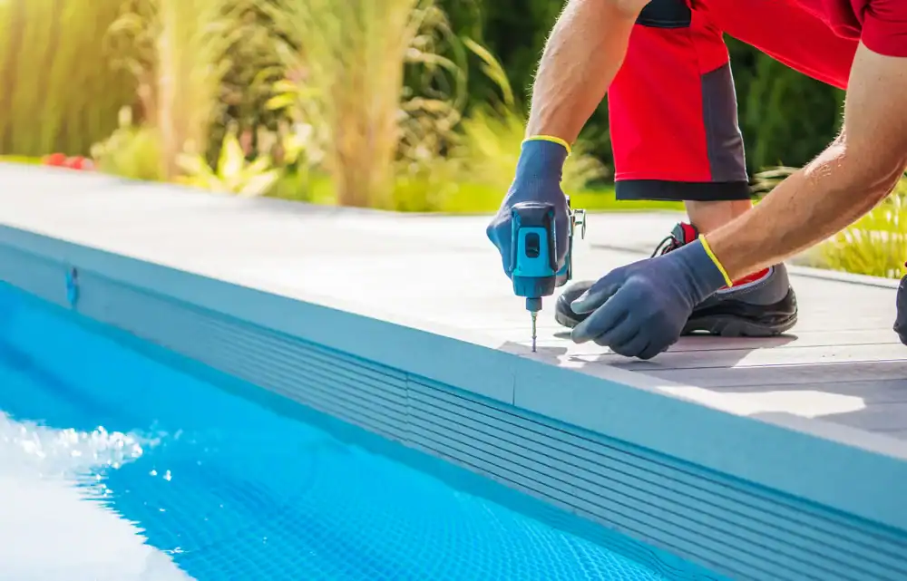 A person in red work pants and gloves uses a power drill to install decking next to a blue swimming pool, showcasing expert pool construction in Douglas County, GA, with green plants in the background.