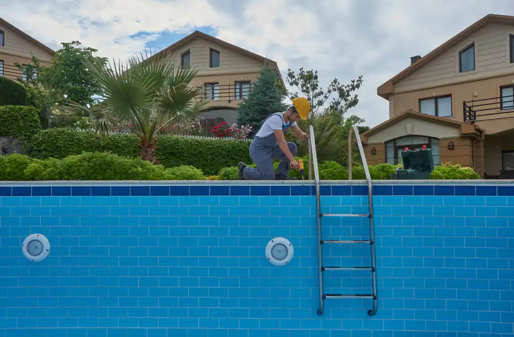 A worker in a yellow hard hat inspects or repairs the ladder of an empty outdoor swimming pool, showcasing pool construction in Douglas County, GA, with houses and greenery in the background.