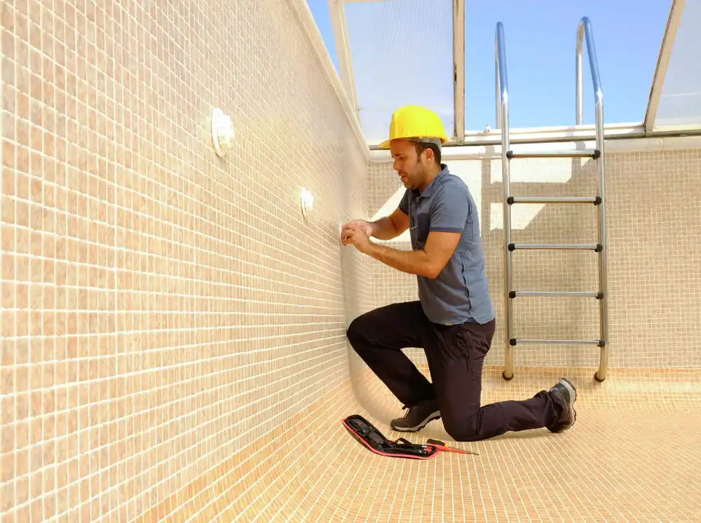 A worker wearing a yellow hard hat kneels inside an empty tiled swimming pool in Douglas County, GA, using tools to inspect or repair a wall fixture near a metal pool ladder under a sunny sky—showcasing expert pool construction.