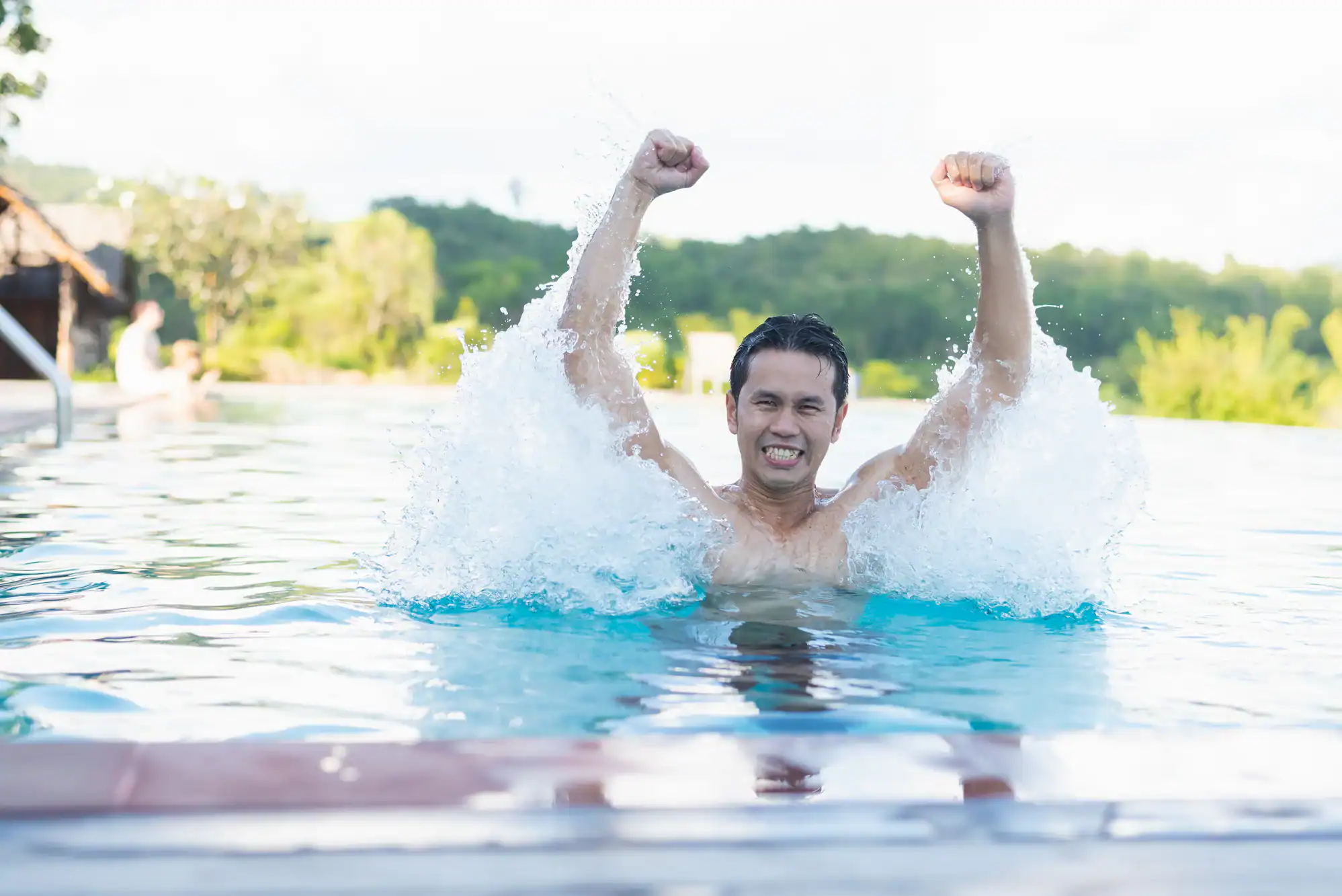 A man in a swimming pool raises his arms triumphantly, splashing water around him, with a big smile and a backdrop of greenery and bright daylight.
