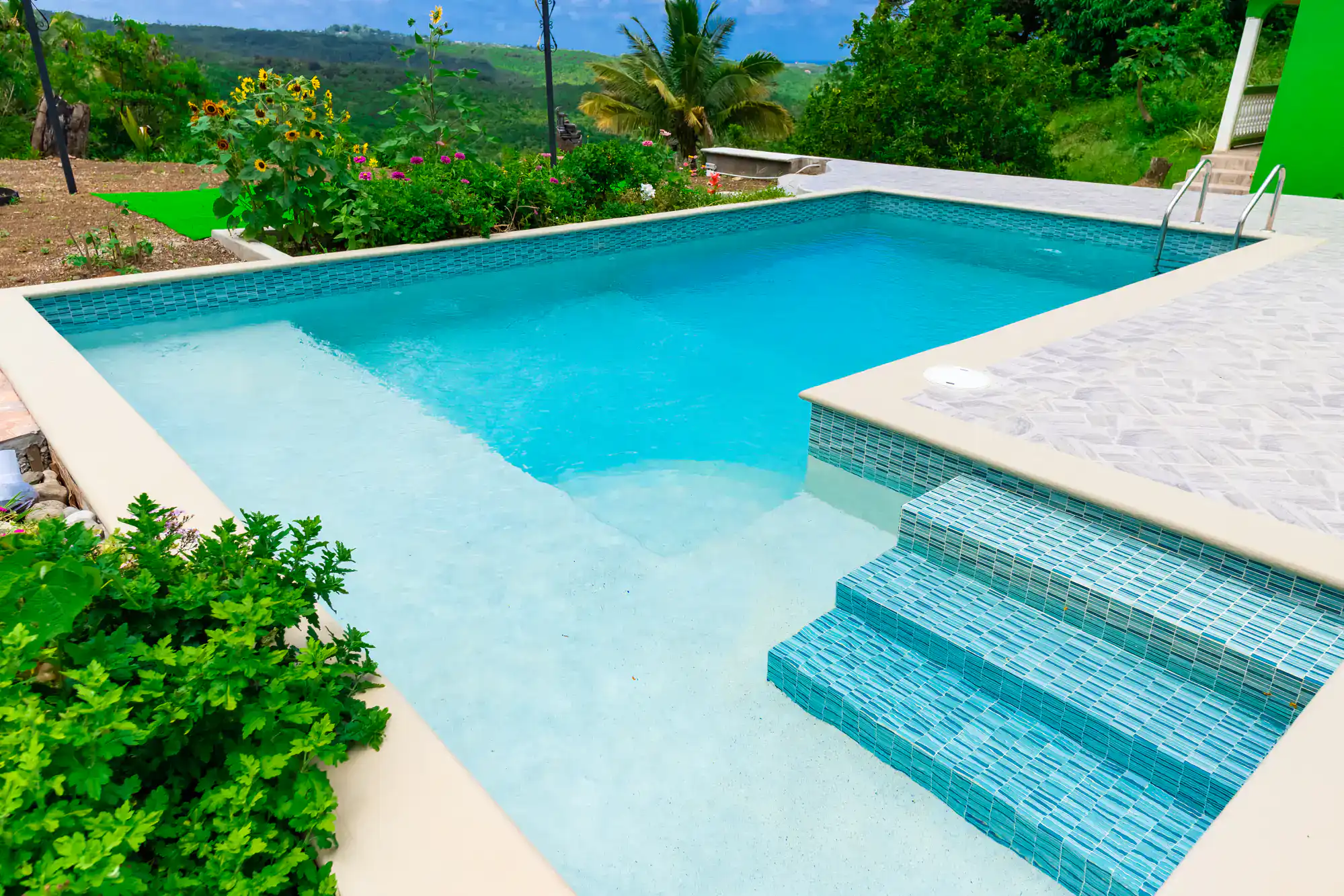 A turquoise swimming pool with tiled steps and a shallow entry overlooks a lush garden with sunflowers, palm trees, and hills in the background under a partly cloudy sky.