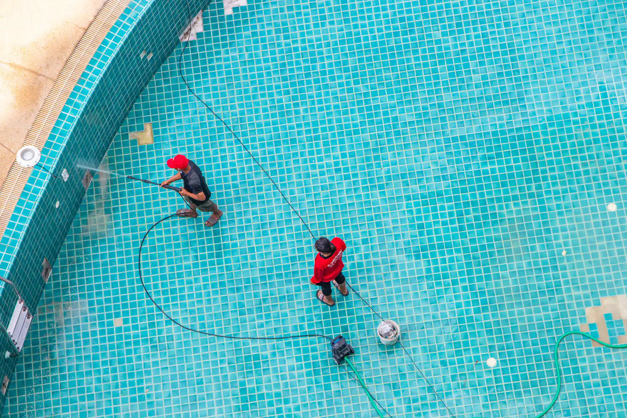 Two workers in red clothing clean an empty swimming pool with turquoise tiles using hoses and cleaning equipment. The scene is viewed from above.