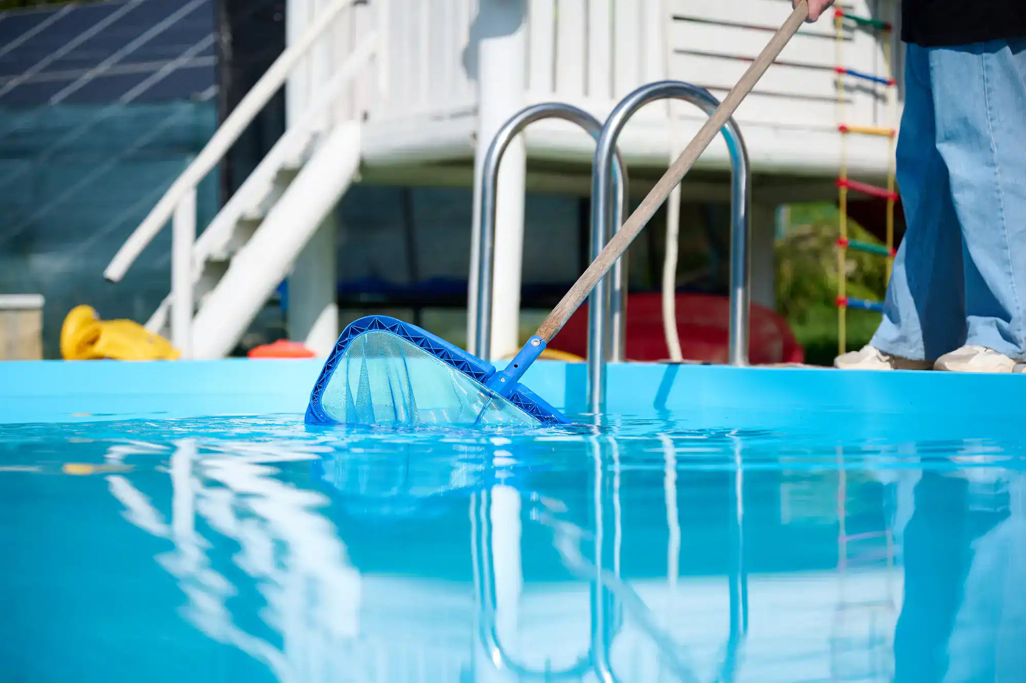 A person uses a blue pool skimmer net on a long pole to clean the surface of an outdoor swimming pool, with a white ladder and building visible in the background.