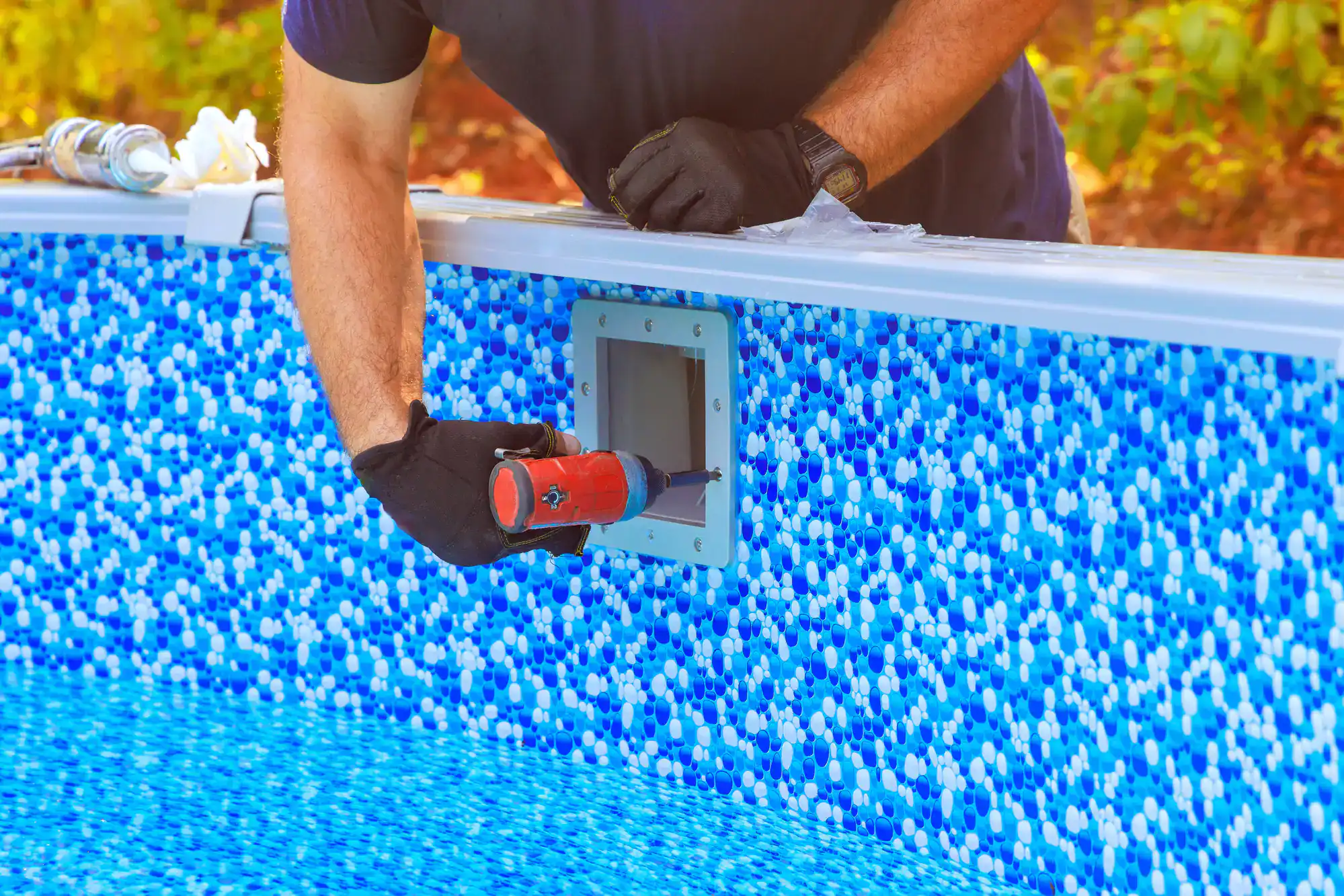 A person wearing black gloves uses a caulking gun to seal a pool skimmer on the blue patterned wall of an above-ground swimming pool.