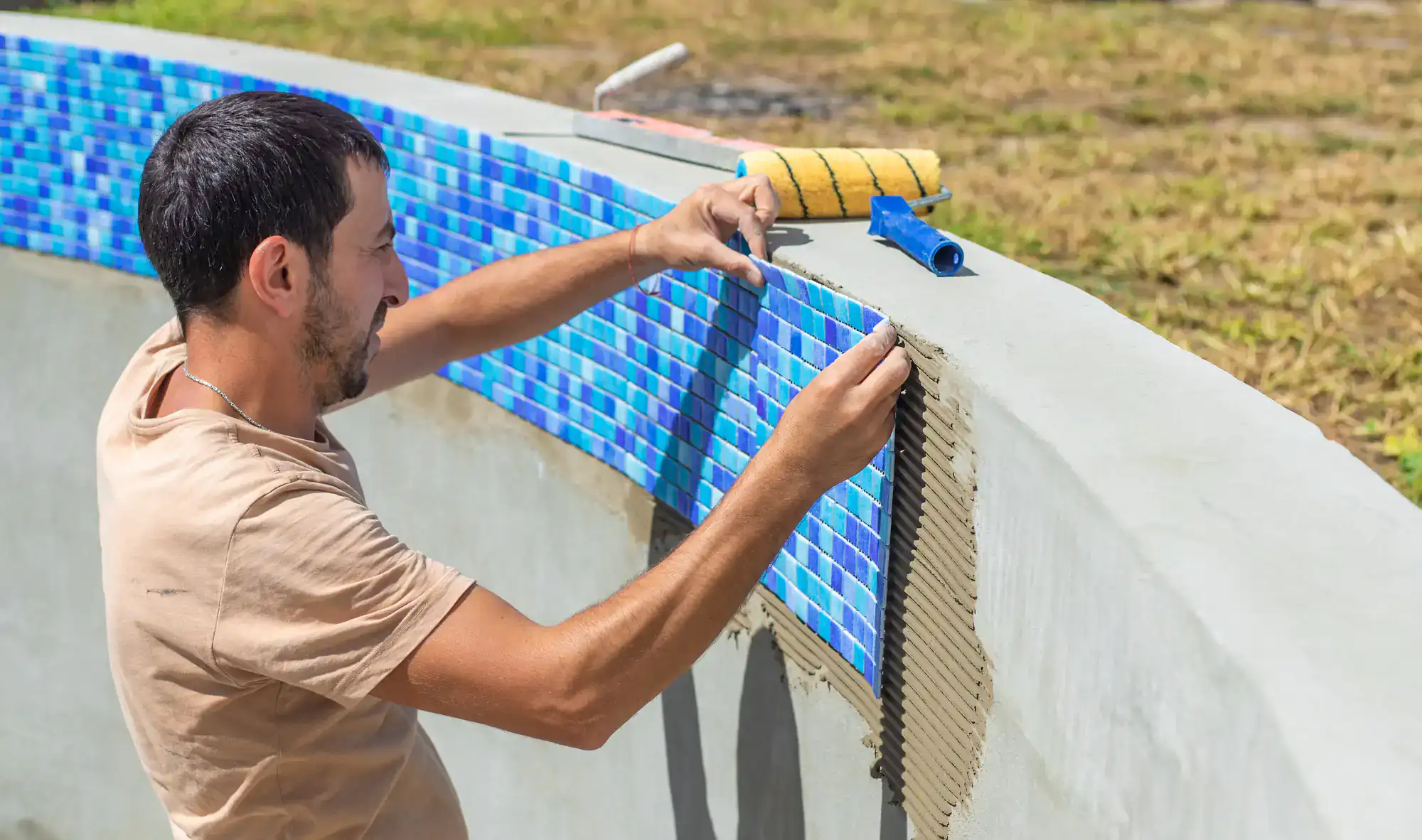 A man installs blue mosaic tiles on the curved inner wall of an empty swimming pool, using adhesive and tools placed nearby, with grass visible in the background.