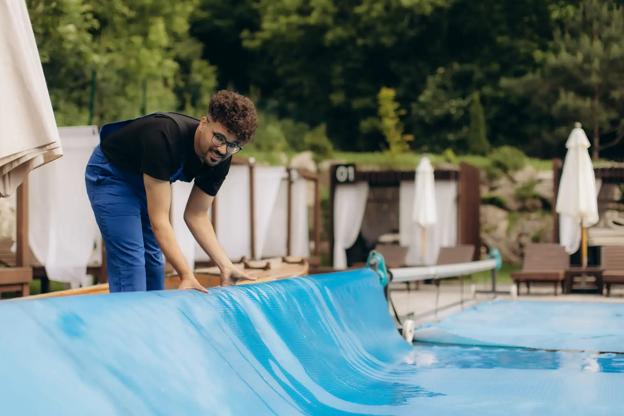 A man in a black shirt and blue pants is unrolling or rolling up a blue pool cover at an outdoor swimming pool, with lounge chairs, umbrellas, and greenery in the background.