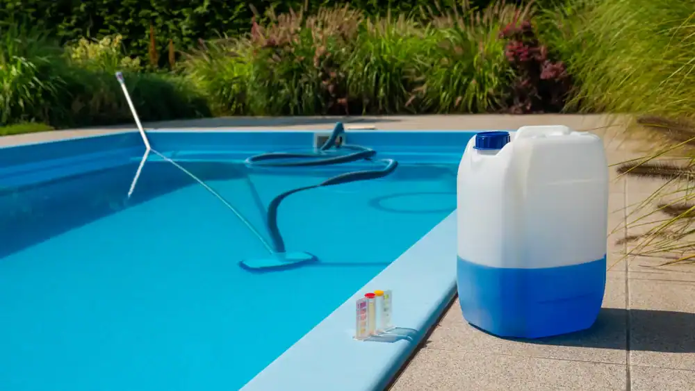 A swimming pool with a cleaning pole and hose in the water, a large plastic chemical container on the poolside, and a small pool water testing kit nearby—showcasing quality Pool Construction Douglas County, GA. Green shrubs and grass surround the area.