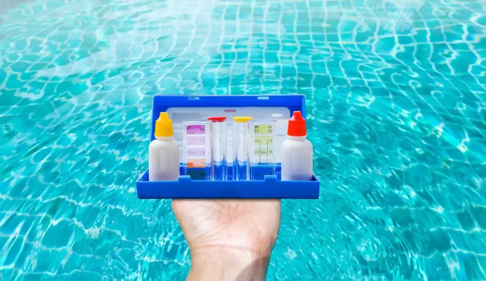 A hand holds a blue plastic pool water testing kit with bottles, test tubes, and color charts, set against the backdrop of a clear swimming pool—perfect for ensuring water quality after Pool Construction in Douglas County, GA.