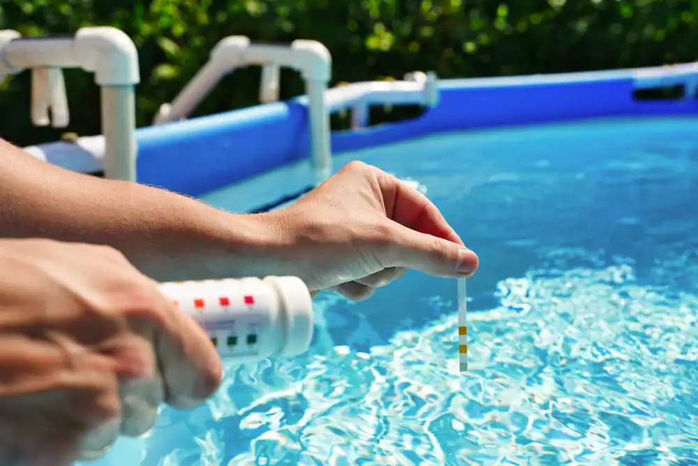 A person’s hand holds a test strip and container above a clear blue pool, checking the water quality—an essential step after Pool Construction in Douglas County, GA. White pool piping and green foliage are visible in the background.