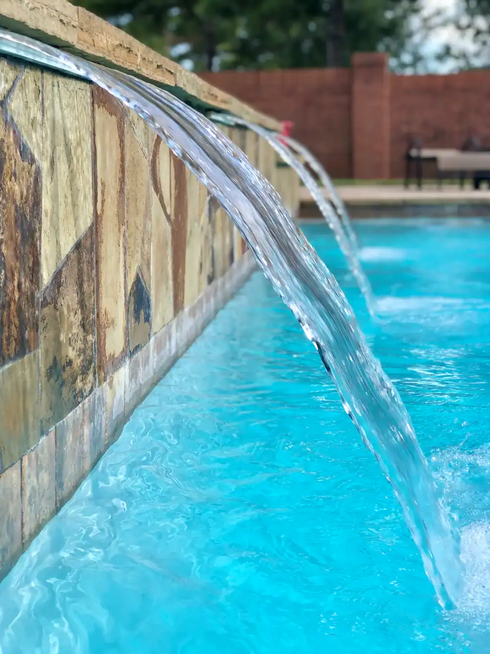 Clear water streams from a decorative stone wall into a bright blue swimming pool, with a blurred background showing a brick fence and outdoor seating.