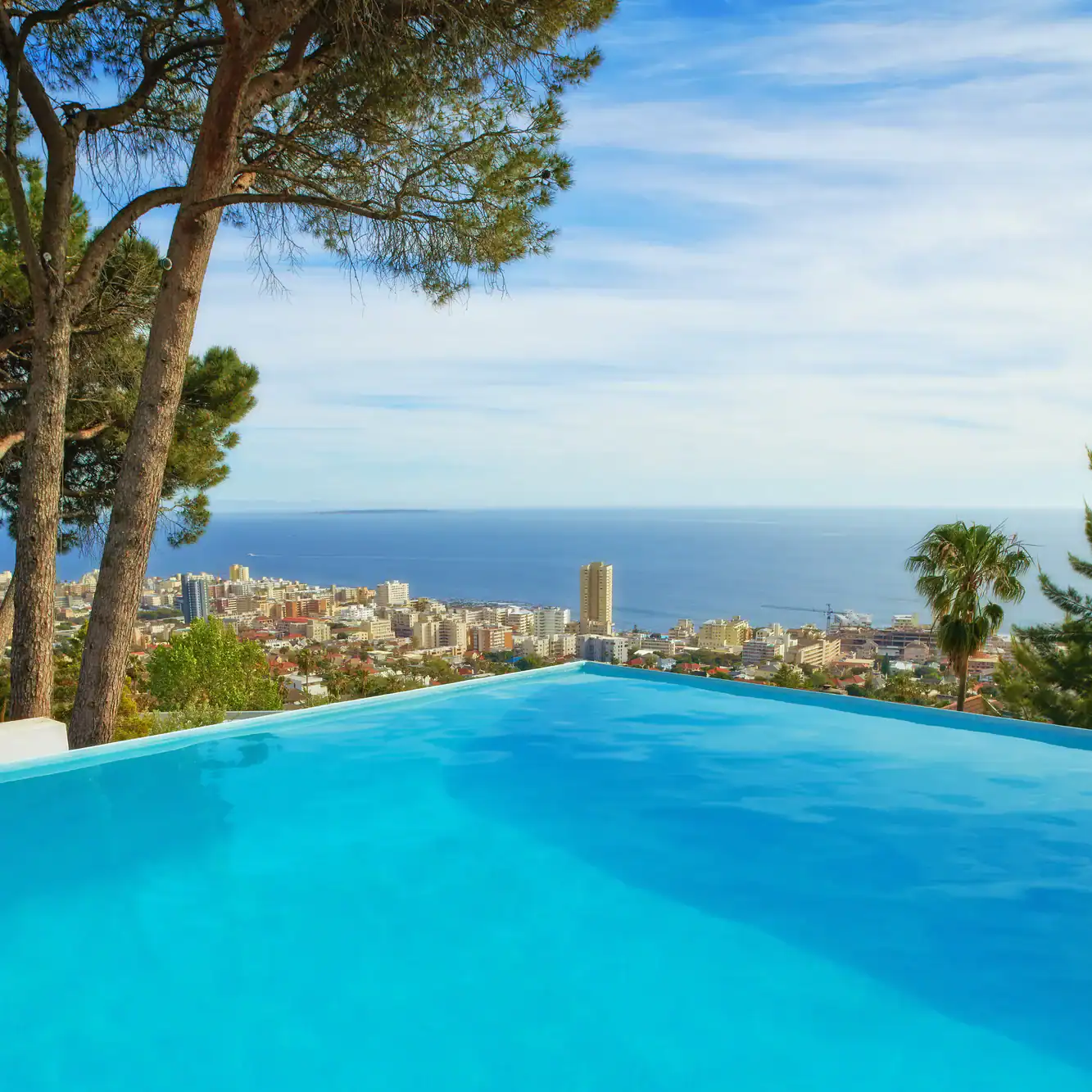 An infinity pool overlooking a coastal city with tall buildings, lush trees, and the blue sea in the distance under a partly cloudy sky.