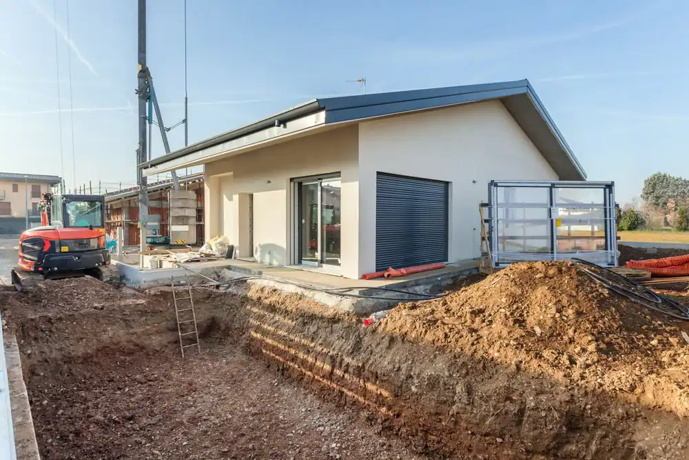 A modern, single-story house under construction with an excavated area and dirt mound in the foreground, construction equipment, and building materials around the site on a clear day.