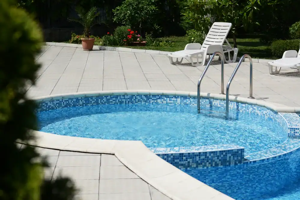 A round swimming pool with clear blue water and metal handrails, surrounded by a tiled deck. White lounge chairs and greenery are visible in the background.