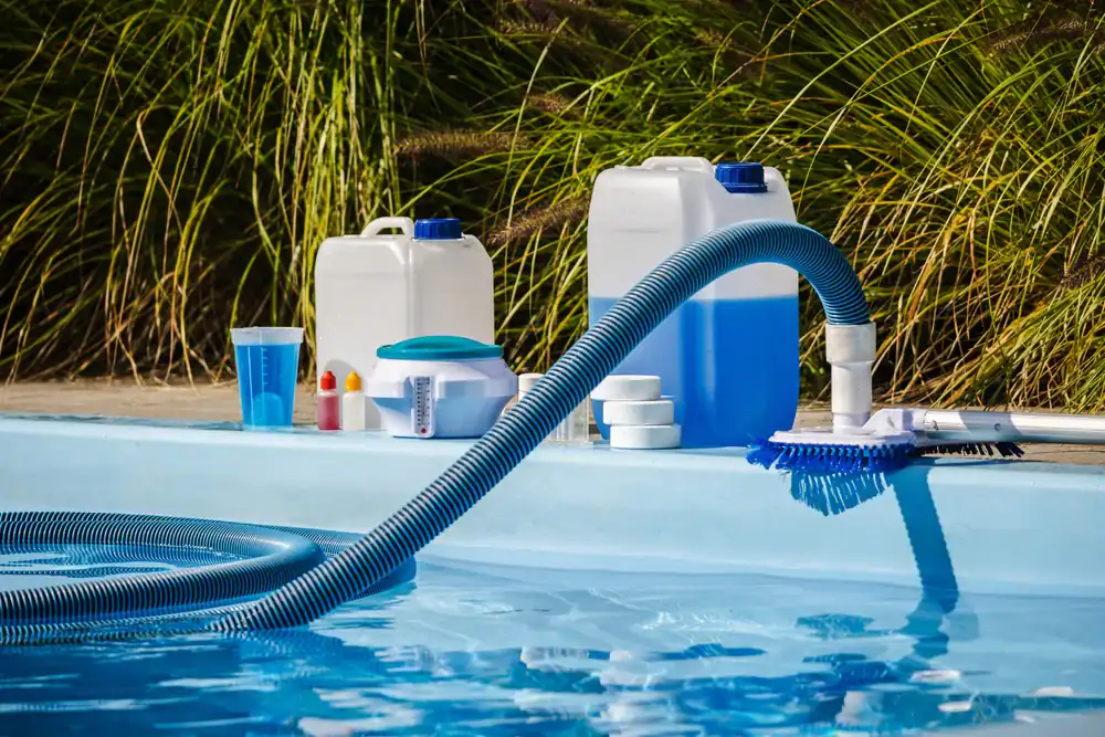A pool cleaning hose and brush in a swimming pool, with large chemical containers, chlorine tablets, testing kits, and cleaning supplies on the poolside, surrounded by tall greenery.