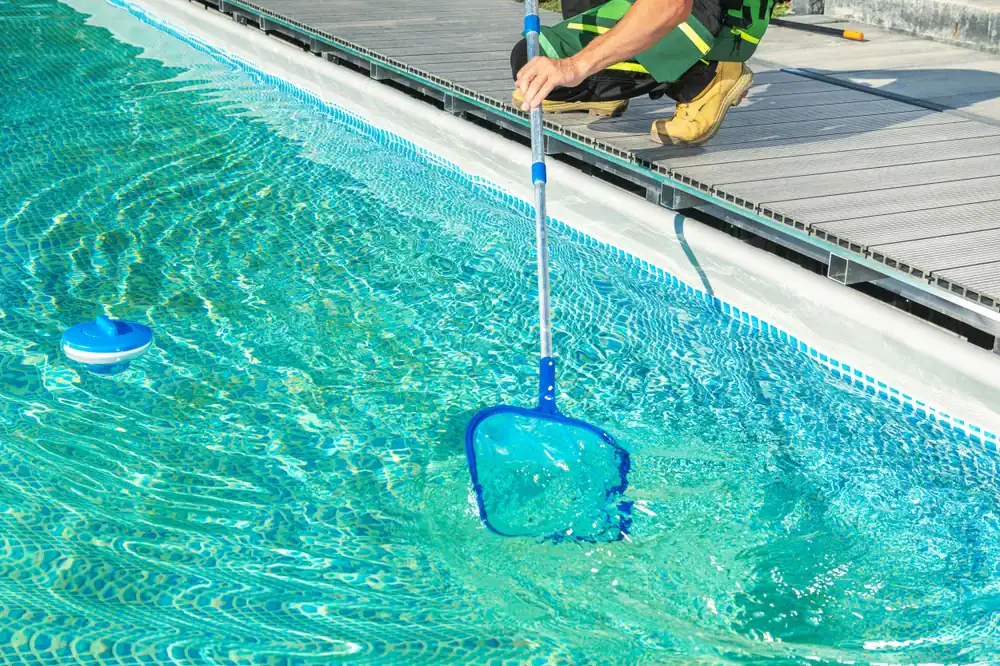A person in work clothes and boots uses a blue pool skimmer net to clean debris from a clear swimming pool near a wooden deck.