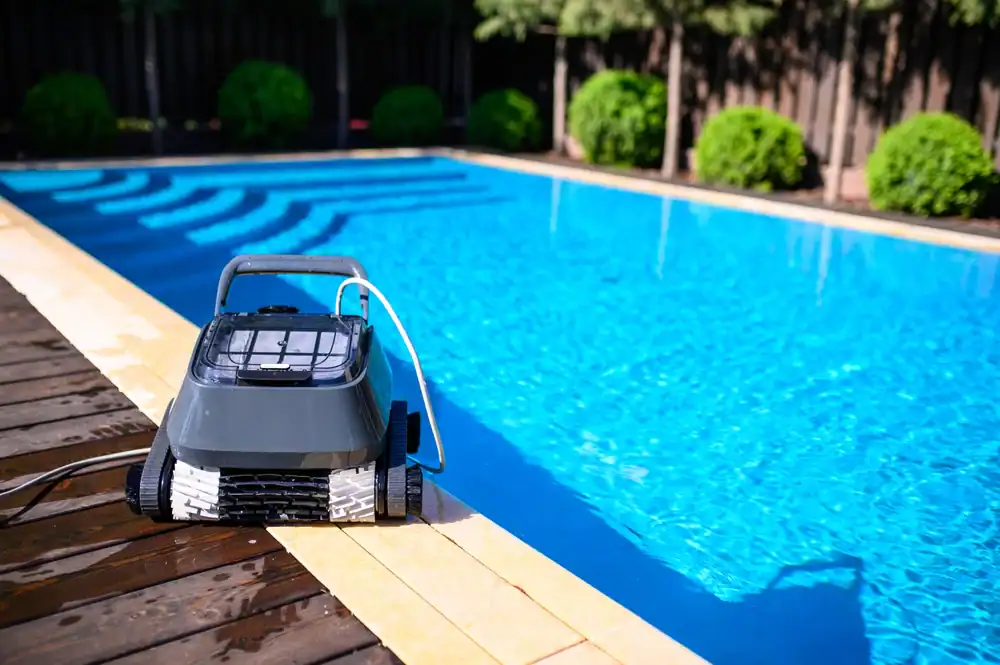 A robotic pool cleaner is positioned on the edge of a bright blue outdoor swimming pool, with trees and bushes in the background.