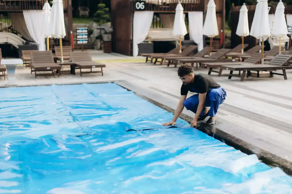 A person in blue pants and a black shirt is kneeling by a pool, adjusting a blue pool cover. Lounge chairs and white umbrellas are visible in the background at a resort or poolside area.