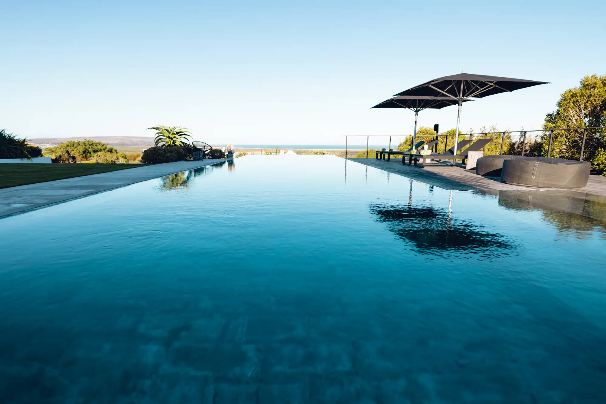 Infinity pool with clear blue water, bordered by a patio with black lounge chairs and a large black umbrella, overlooking greenery and a distant horizon under a clear sky.