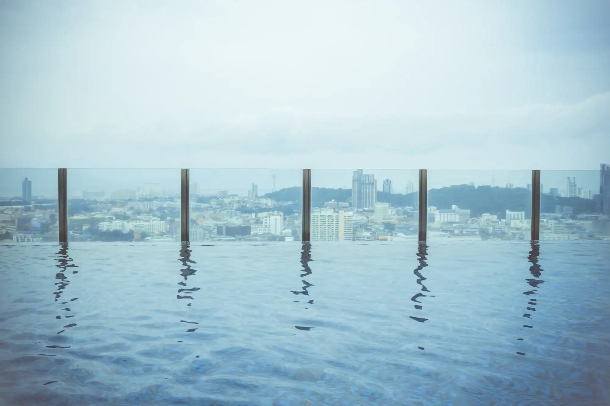 A rooftop infinity pool with calm water overlooks a cityscape through a glass barrier. The skyline is slightly hazy under a cloudy sky, with buildings and distant hills visible in the background.