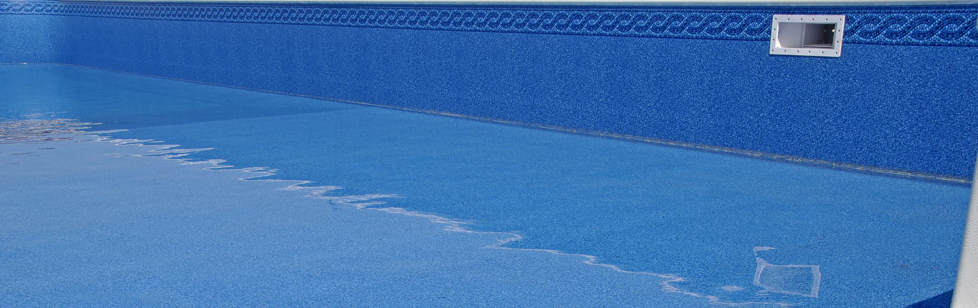 A swimming pool partially filled with clean blue water, showing the underwater slope from the shallow end to the deep end, with a patterned tile border along the top edge of the pool wall.