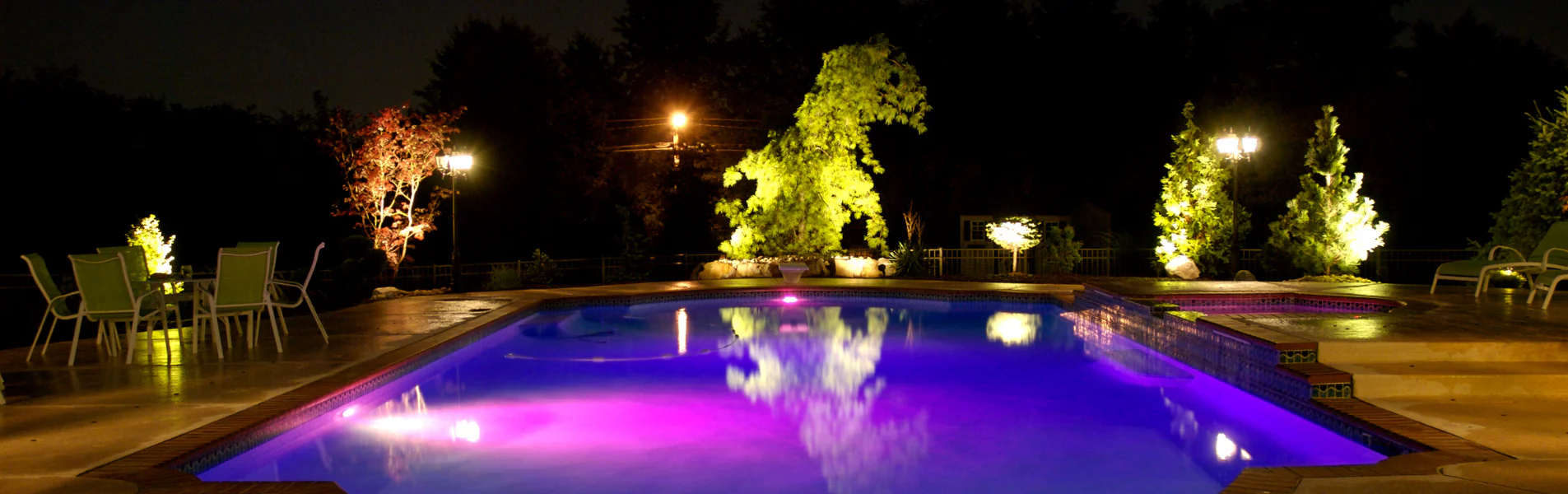 A swimming pool at night, illuminated with purple lights, surrounded by trees and plants lit by warm lamps, with patio chairs arranged on the poolside.