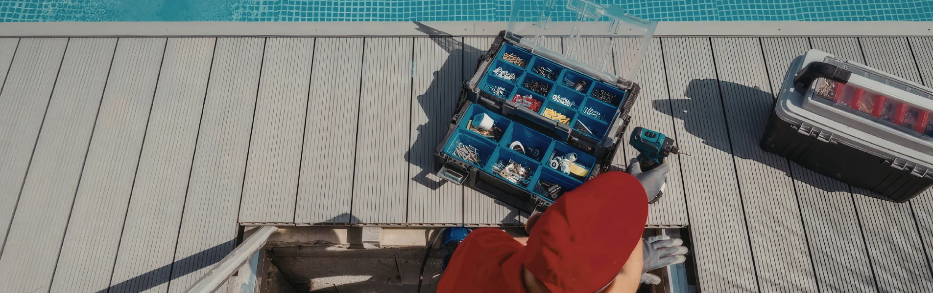 A person in a red shirt and gloves kneels by an open toolbox filled with various screws and tools next to a pool, preparing for maintenance or repair work.