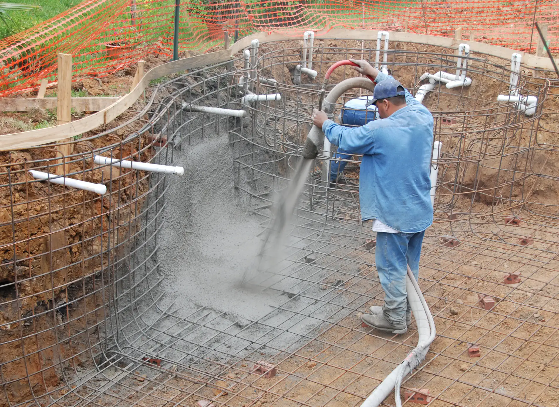 A construction worker in blue clothing sprays concrete onto a rebar framework, forming the curved wall of an in-ground pool at a construction site surrounded by dirt and orange safety fencing.
