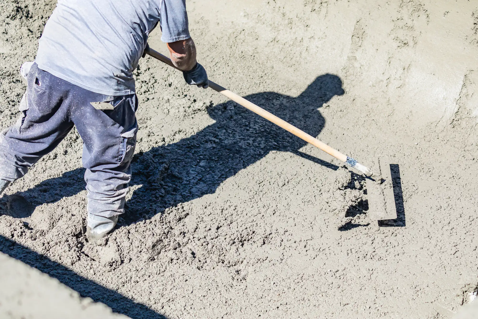 A construction worker wearing boots and gloves uses a long-handled tool to smooth and level wet concrete on a construction site.