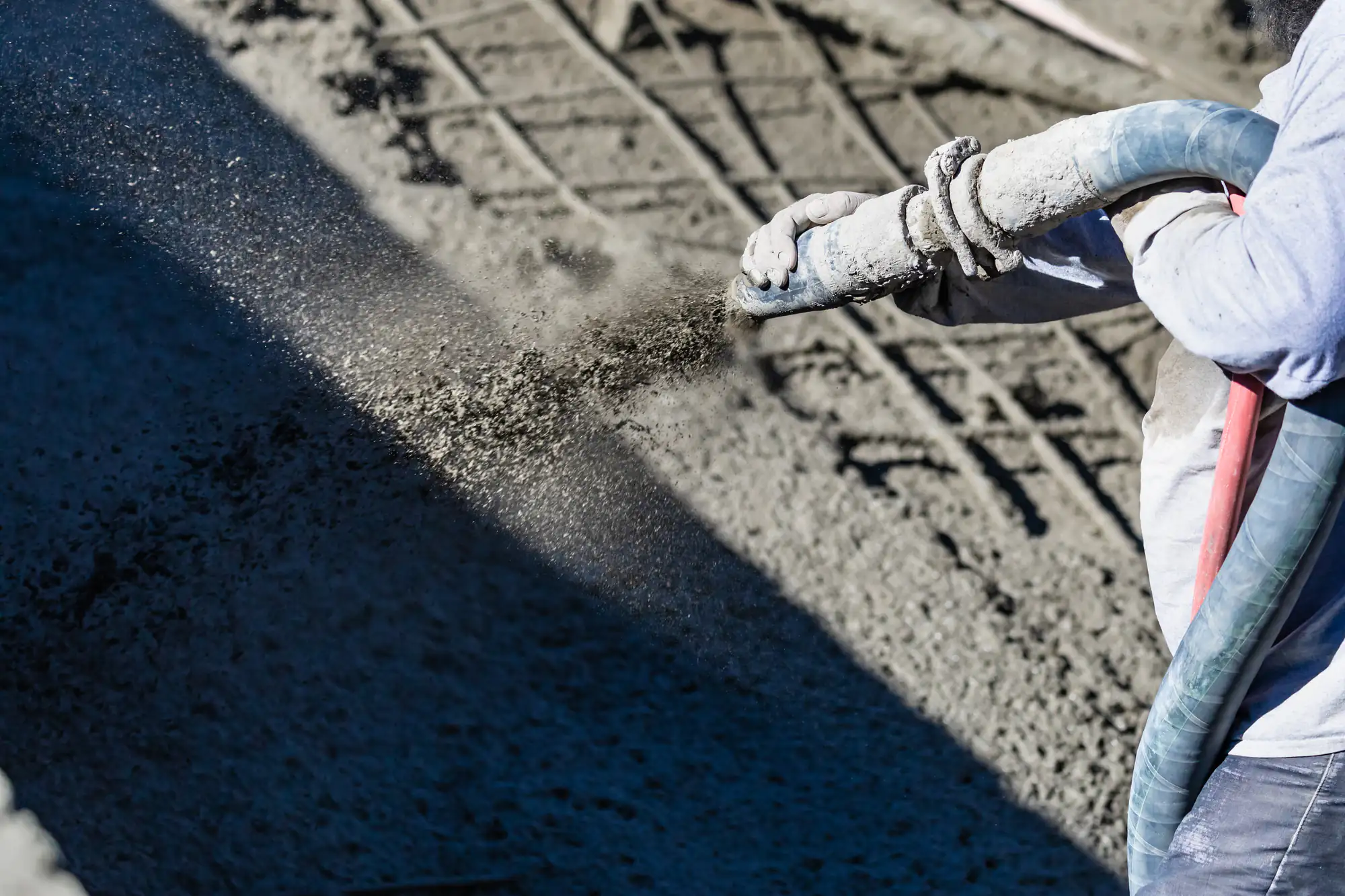 A construction worker sprays concrete onto a surface using a hose, applying a layer of wet concrete for building or repair work. Only the worker's arm and part of the hose are visible.