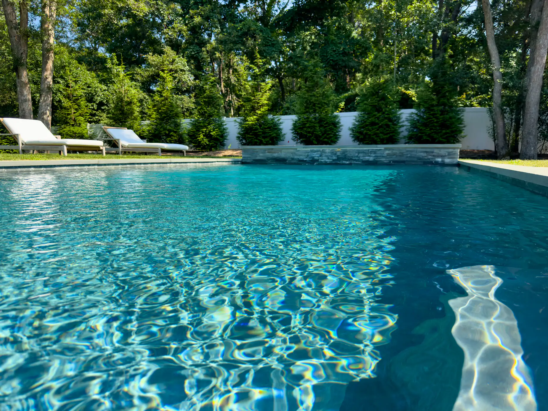A clear blue swimming pool surrounded by green trees and bushes, with white lounge chairs on a grassy area in the background under bright sunlight.
