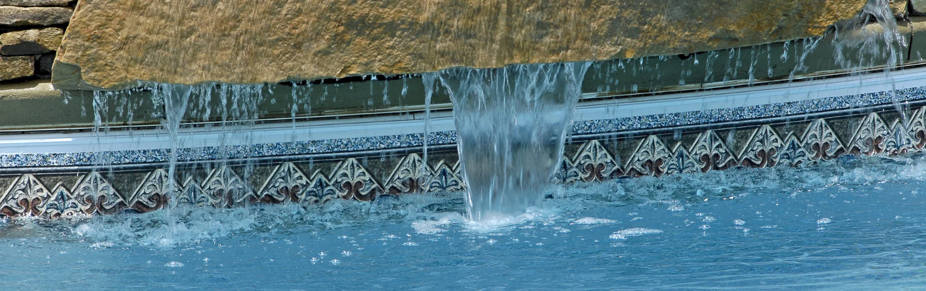 Water cascades over a stone ledge into a swimming pool, creating small streams and splashes. Decorative tile with a geometric pattern lines the pool’s edge.