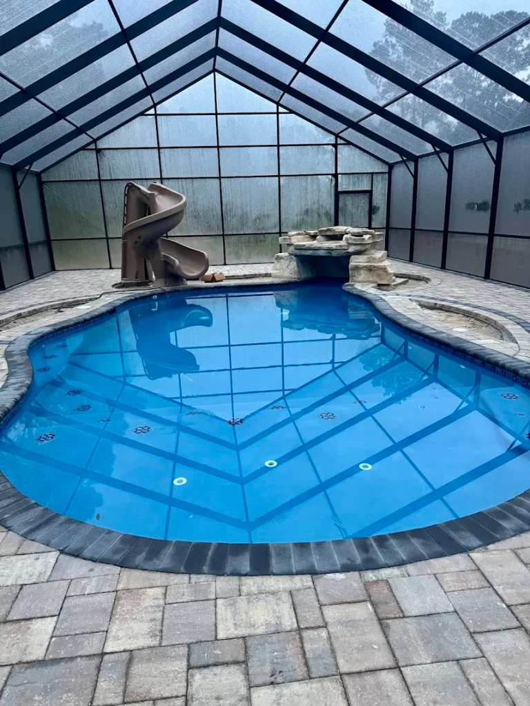 Enclosed pool with water slide, rock waterfall, and paved deck inside a screened structure for relaxation.